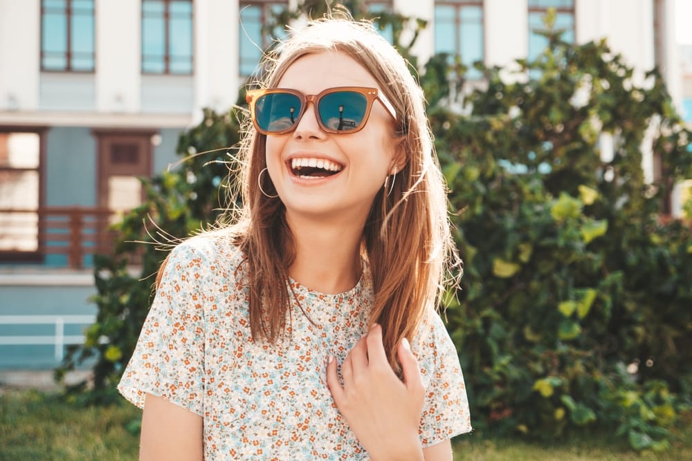 A smiling woman outdoors wearing orange-framed sunglasses and a floral top, standing in front of green bushes and a building - Yucaipa Optometry Happy woman wearing sunglasses outdoors, showcasing stylish eyewear from Yucaipa Optometry